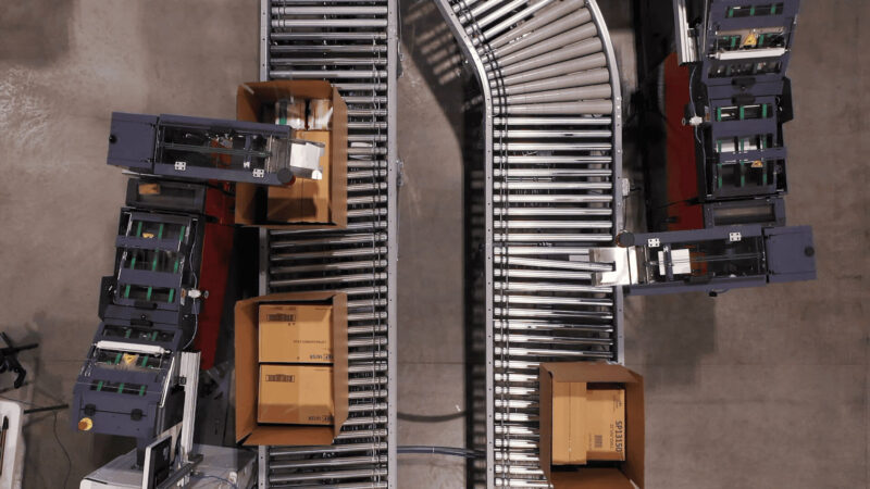 Aerial view of a warehouse conveyor system with two parallel roller conveyors curving to the right, automated sorting machines in dark gray and purple on the left, and scattered cardboard boxes throughout the facility.