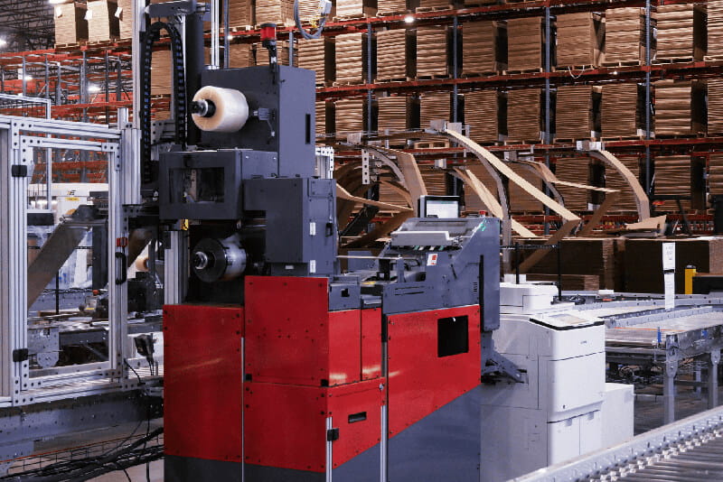 Dark gray and red automated packaging machine with plastic wrap, cardboard processing, and shelving in the background.