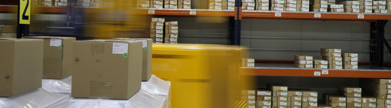 A long-exposure warehouse shot blurs a yellow forklift and cart in motion, with cardboard boxes on white pallets in the foreground and tall dark-metal shelving with orange crossbeams stacked with boxes on the right.