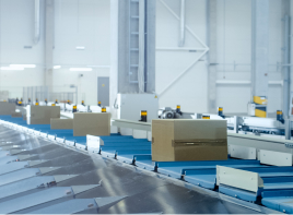 A clean, modern automated warehouse features a blue conveyor belt carrying light brown cardboard boxes diagonally across the frame, with white machinery and small yellow sensors in the background.