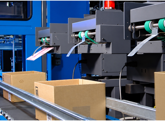 Empty cardboard boxes move along a conveyor beneath dark gray labeling or printing machines with green rollers feeding striped paper, set against blue industrial machinery in a well-lit facility.
