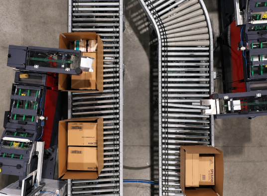 An aerial view shows an industrial roller conveyor curving through a warehouse, transporting three cardboard boxes with smaller red and gray feeder belts merging into the main line on a light gray concrete floor.