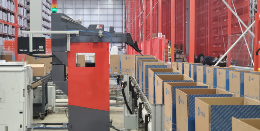 A long shot of an automated warehouse shows rows of uniform open boxes on a conveyor beside a large red-and-black robotic arm, backed by towering red shelving units.