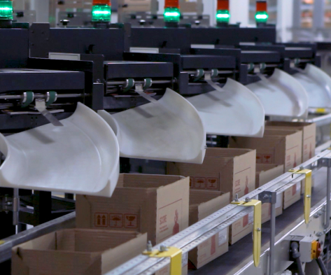 A close-up of an automated sorting system shows cardboard boxes moving under black mechanical arms with white scoop-like attachments, each topped with a green indicator light.