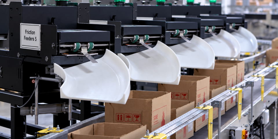 A row of five black-and-white Friction Feeders with curved white chutes stands above a conveyor belt carrying open cardboard boxes marked with red and black shipping symbols in an industrial setting.