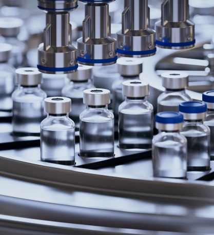 A production line of small glass vials filled with clear liquid moves along a conveyor beneath robotic filling nozzles.