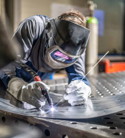 A welder in a protective helmet and gloves bends over a metal sheet, torch sparking brightly as they weld.